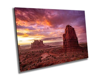 May include: A vibrant sunset over a desert landscape with towering red rock formations. The sky is a mix of pink, purple, and orange hues, with clouds casting long shadows across the scene.