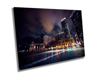 May include: A night view of a city skyline with tall buildings illuminated by streetlights. The street is wet from rain and reflects the city lights.