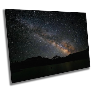 Puede incluir: Una impresión rectangular del cielo nocturno con la Vía Láctea. La imagen muestra un cielo oscuro y estrellado con una brillante banda de estrellas y colores. Montañas silueteadas y un lago en primer plano.