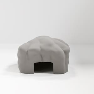 Può includere: Un oggetto decorativo grigio a forma di grotta. L'oggetto ha un'apertura rettangolare e una superficie strutturata a forma di roccia. La grotta è di colore grigio chiaro e poggia su una superficie bianca.