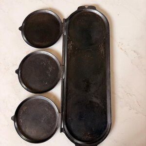 Pode incluir: Uma chapa de ferro fundido preto com três tampas circulares articuladas presas a uma base retangular longa. A base apresenta três reentrâncias circulares e cada tampa tem uma pequena aba lateral para abertura. Superfície metálica com desgaste.