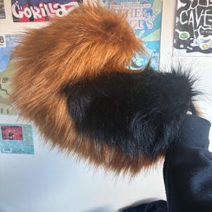 May include: A plush, curved tail accessory made of long, shaggy faux fur. The tail features a distinct colour-blocked design with a vibrant, warm orange section at the top and a deep, solid black section at the base, creating a two-tone appearance.