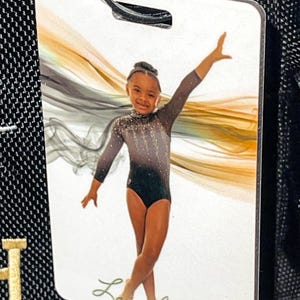 May include: A white luggage tag featuring a young gymnast in a black and gray leotard with a gold and black abstract design. The tag has the name "Layiah Perry" in cursive.