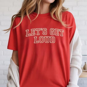 May include: A bright red short-sleeved t-shirt featuring the text "LET'S GET LOUD" printed in a bold, white, collegiate-style font with a thin yellow outline across the center of the chest. The shirt has a crew neckline and a relaxed fit.