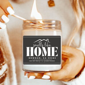 May include: A white candle with a black label that reads "smells like HOME NEWNAN, GA 30263 100% SOY WAX 9 oz HAND POURED". The candle is lit and the flame is visible.