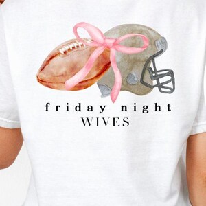 May include: White t-shirt featuring a watercolor design of a football and helmet tied with a pink bow. The text "friday night wives" is printed below the graphic. The shirt is made of a soft material.