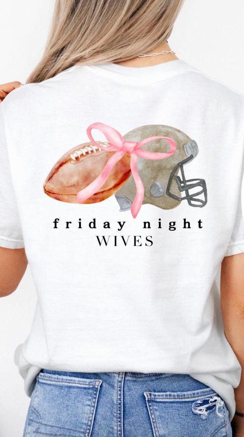 May include: White t-shirt featuring a watercolor design of a football and helmet tied with a pink bow. The text "friday night wives" is printed below the graphic. The shirt is made of a soft material.