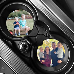 May include: Black car cup holder with two circular inserts, each displaying a family photo. One insert shows two young boys, and the other shows a family of four. The holder is made of black plastic.