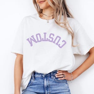 May include: White t-shirt with the word "CUSTOM" printed in a lavender color across the chest. The shirt is a crew neck style and appears to be made of a soft material. The model is wearing blue jeans.