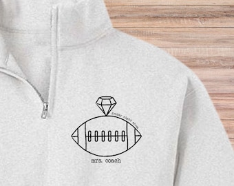 Peut inclure: Un sweat-shirt gris clair à fermeture éclair quart de zip avec un motif noir d'un ballon de football surmonté d'un diamant. Le texte "friday night wives" et "mrs. coach" est en noir.
