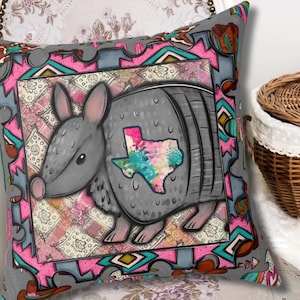 May include: Gray decorative pillow featuring a cartoon armadillo with a Texas outline. The pillow has a pink, turquoise, and gray border with a patterned background. The armadillo has a colorful Texas outline on its side.