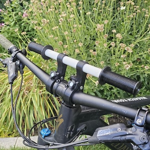 May include: A mountain bike handlebar with black grips and a white bar. The bike has a black frame and blue suspension controls. The background shows green foliage and flowers.