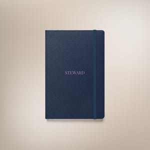 May include: A navy blue journal with the word "STEWARD" printed in a lighter shade of blue. The journal has a matching elastic band closure. The cover has a textured appearance.