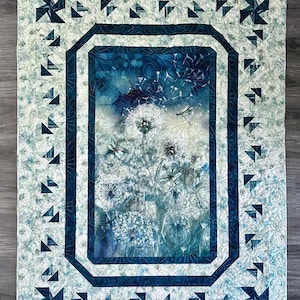 May include: A quilted wall hanging featuring a central image of dandelions and dragonflies in shades of blue and white. The quilt has a dark blue border and a light blue patterned background with geometric accents. The overall theme is nature.