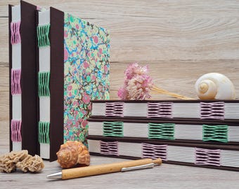 May include: Handmade journals with brown covers and colorful marbled paper. The journals are bound with pink and green stitching. A wooden pen, seashells, and dried flowers are arranged nearby on a wooden surface.