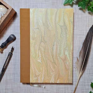 Könnte beinhalten: Ein braunes, ledergebundenes Tagebuch mit einem marmorierten Papierumschlag in Creme-, Gelb- und Grüntönen. Das Tagebuch hat ein braunes Band als Lesezeichen.