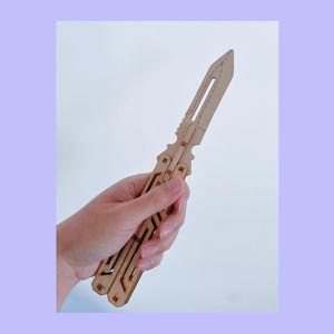 May include: A wooden butterfly knife with a light brown finish. The knife features a blade with a cutout design and a handle with geometric cutouts. The knife is held in a hand against a white and lavender background.