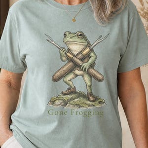May include: A sage green short-sleeve t-shirt featuring a graphic illustration of a frog standing on a rock while holding two crossed fishing rods. Below the frog, the text Gone Frogging is printed in a dark green font on the front of the shirt.