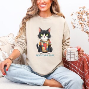 May include: Beige sweatshirt featuring a black and white cat wearing a colorful patchwork sweater. The cat has green eyes and is sitting. The text "SEW OVER THIS" is printed below the cat. The sweatshirt is worn by a person.