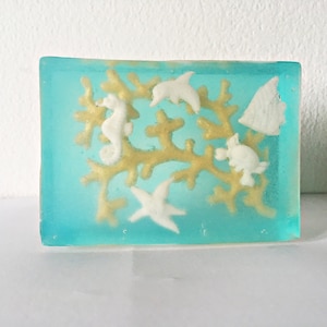 May include: A rectangular bar of soap with a turquoise blue hue, featuring white sea creatures and gold coral. The soap includes a seahorse, starfish, dolphin, turtle, and fish. The soap is a decorative item.
