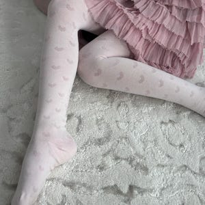 May include: White tights with a delicate pattern of small hearts. The tights are worn by a person sitting on a fluffy white rug.