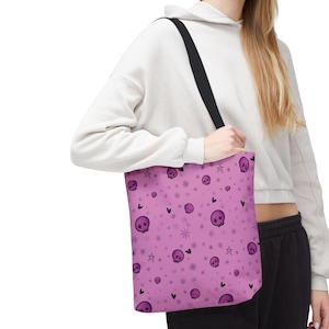 Peut inclure: Un sac fourre-tout rose avec un motif répété de crânes violets, d'étoiles, de cœurs et de flocons de neige. Le sac a une bandoulière noire. La personne porte un sweat à capuche blanc et un pantalon de survêtement noir.