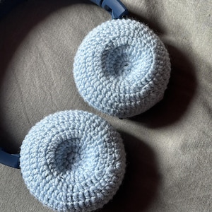 Puede incluir: Un par de fundas de orejeras de auriculares de color azul claro, hechas a ganchillo, descansan sobre una superficie gris. Las fundas de las orejeras son circulares y tienen un aspecto texturizado y hecho a mano. Los auriculares son de color azul oscuro.