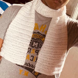 Peut inclure: Une écharpe crochetée blanche à texture côtelée portée sur un t-shirt gris à manches courtes. Le t-shirt présente un motif graphique avec le texte Marshall, la lettre M et le chiffre 83 en lettres jaunes et noires sur le devant.