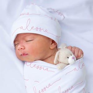 May include: A white swaddle blanket with the name "Alessia" written in pink script. A baby is sleeping in the swaddle, wearing a white hat with the same name in pink script. A small knitted toy is tucked into the swaddle.