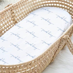 May include: A white baby bassinet sheet featuring the name "Jacob Henry" in a repeating, navy blue cursive script. The sheet is placed inside a woven wicker bassinet.