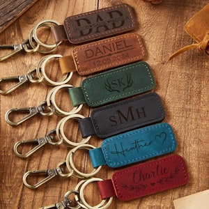 Puede incluir: Un conjunto de siete llaveros de cuero con diferentes nombres y fechas grabados en ellos. Los llaveros son de color marrón, verde, negro, azul y rojo. Los llaveros están colgados en una superficie de madera.