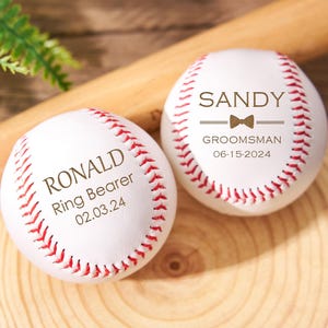 Puede incluir: Dos pelotas de béisbol blancas con costuras rojas y texto personalizado. Una dice "RONALD Ring Bearer 02.03.24" y la otra dice "SANDY GROOMSMAN 06-15-2024" con un lazo. Un bate de béisbol de madera y una rodaja de madera están en el fondo.