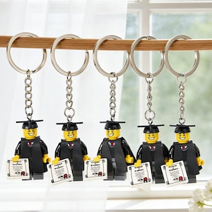 May include: Five keychains featuring graduation-themed Lego figures. Each figure wears a black graduation gown and mortarboard, holding a diploma. The figures have yellow heads and are attached to silver keychains.