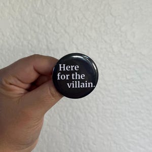 Op de afbeelding: Een zwarte knoop met de witte tekst "Here for the villain". De ronde knoop heeft een glanzend oppervlak. De knoop wordt in een hand gehouden tegen een witte achtergrond.