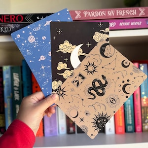 May include: Three decorative notebooks with celestial designs. One is blue with planets and stars, another is black with a moon and clouds, and the third is tan with suns, moons, snakes, and constellations. The notebooks are held in front of a bookshelf.