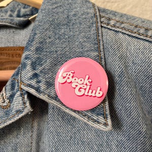 May include: A round, pink button with the words "Book Club" in white script and red outline. The button is pinned to a light blue denim jacket.