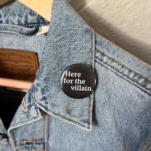 Op de afbeelding: Een lichtblauwe spijkerjas met een zwarte knoop op de kraag gespeld. De knop heeft witte tekst met de tekst "Here for the villain."