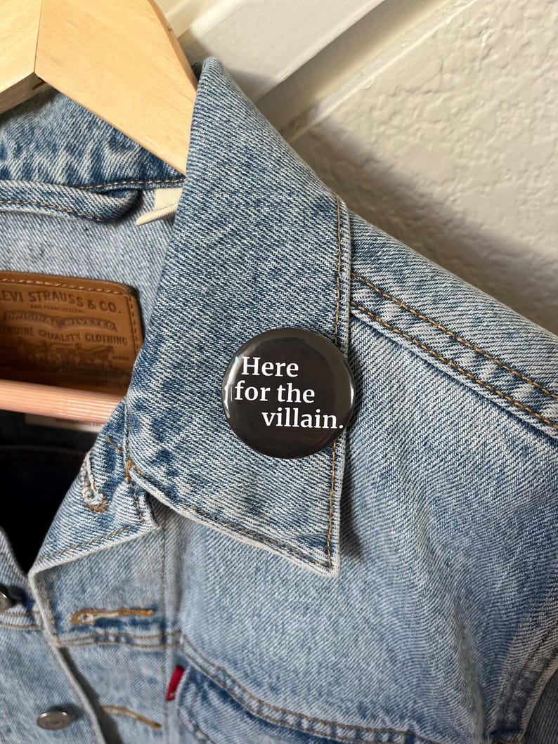 Op de afbeelding: Een lichtblauwe spijkerjas met een zwarte knoop op de kraag gespeld. De knop heeft witte tekst met de tekst "Here for the villain."