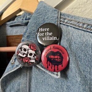 Op de afbeelding: Drie ronde buttons op een spijkerjasje. E&eacute;n button is zwart met de tekst "Here for the villain.", een andere is grijs met twee schedels en rozen, en de derde is rood met druipende lippen en de woorden "Dark Romance."