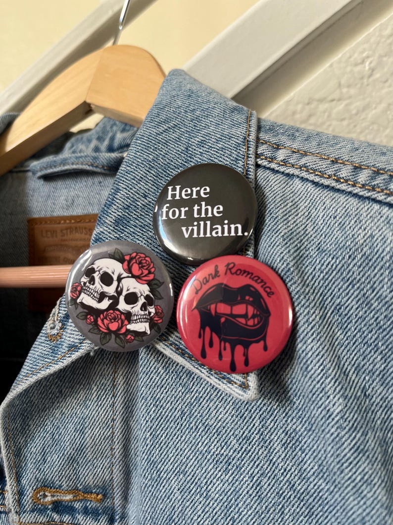 Op de afbeelding: Drie ronde buttons op een spijkerjasje. E&eacute;n button is zwart met de tekst "Here for the villain.", een andere is grijs met twee schedels en rozen, en de derde is rood met druipende lippen en de woorden "Dark Romance."