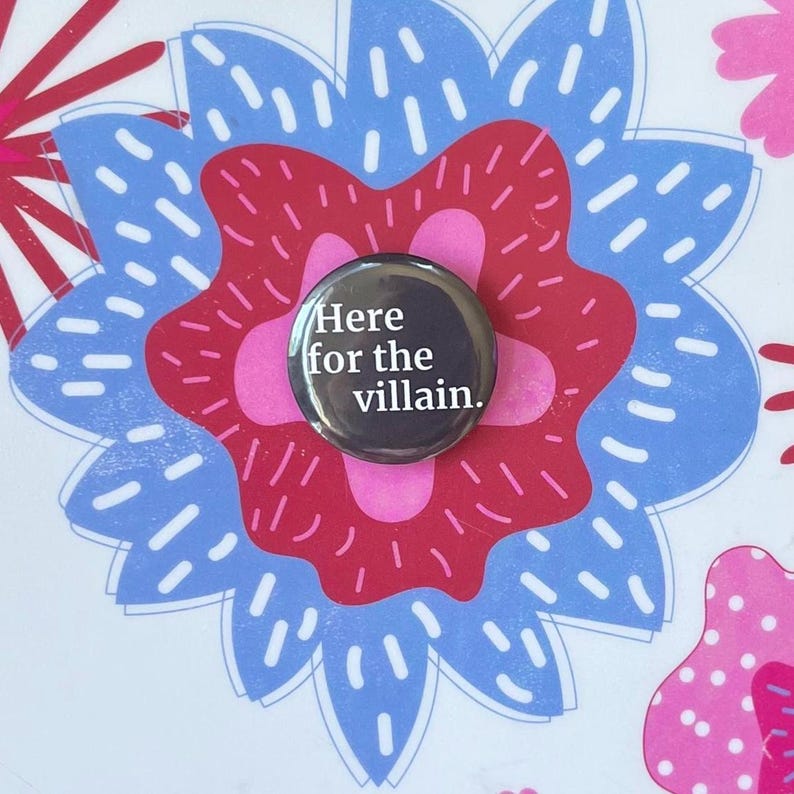 Op de afbeelding: Een ronde, zwarte button met de tekst "Here for the villain." in witte letters. De button is geplaatst tegen een bloemmotief in roze, rood en blauw.