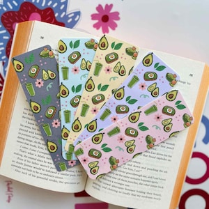 May include: A collection of colorful bookmarks with avocado and smoothie illustrations. The bookmarks are on top of an open book. The bookmarks have different background colors, including gray, blue, yellow, and pink.