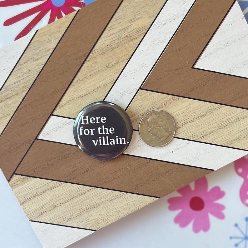 Op de afbeelding: Een ronde, zwarte button met de tekst "Here for the villain." in witte letters. Een munt ligt naast de button. De button en de munt liggen op een houten oppervlak met een geometrisch patroon.