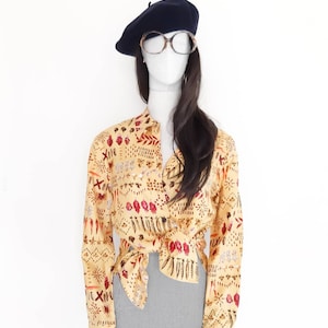 May include: A yellow long-sleeved shirt with a colorful pattern of flowers, leaves, and geometric shapes. The shirt is tied at the waist and paired with a gray skirt.