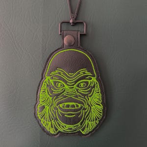 May include: A black keychain with a green embroidered creature design. The keychain has a metal clasp and a leather-like backing. The creature's face is detailed with green stitching, and the overall design is a unique accessory.