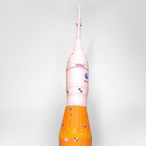 May include: A model of a space shuttle with three orange and white rocket boosters. The shuttle has a white nose cone with the NASA logo and the text "NASA" printed on it.