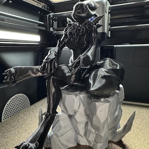 May include: A 3D printed black figurine of a creature with a long tail and claws, sitting on a gray rock base. The figurine is detailed and has a textured surface.