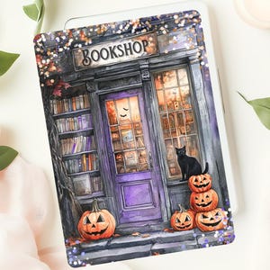 May include: A decorative tablet cover featuring a whimsical illustration of a "BOOKSHOP" with a purple door, pumpkins, and a black cat. The design has a Halloween theme with a cozy, inviting atmosphere, perfect for book lovers.