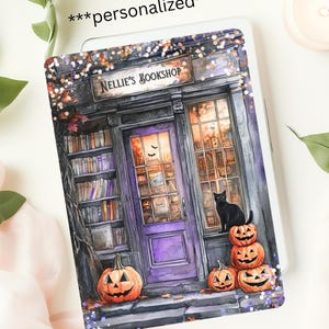 May include: A personalized tablet cover featuring a watercolor illustration of a spooky bookstore. The cover displays a purple door, pumpkins, a black cat, and the text "NELLIE'S BOOKSHOP". The design has a Halloween theme.