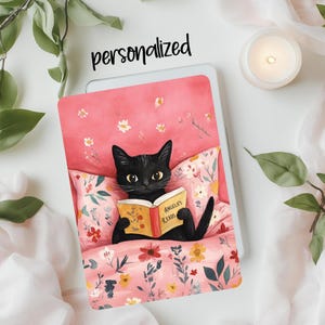May include: A black cat with yellow eyes is lying in a pink bed with a floral pattern. The cat is holding a red book with the words "Angela's Reads" on the cover.